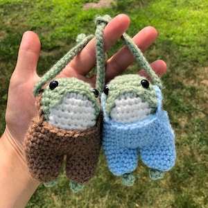 Puede incluir: Dos peluches de rana de crochet con cuerpos verdes, caras blancas y ojos negros. Una rana lleva un mono marrón y la otra lleva un mono azul.