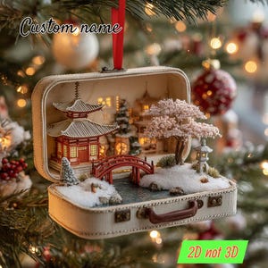 Puede incluir: Diorama en miniatura de una escena japonesa dentro de una pequeña maleta. La escena incluye un puente rojo, una pagoda y un cerezo en flor. El texto "Custom name" está encima de la maleta y "2D not 3D" debajo.