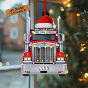 May include: Red semi-truck Christmas ornament with a Santa hat and colourful lights. The ornament has the name "John" on the front and the text "THIS ORNAMENT IS FLAT, NOT 3D".