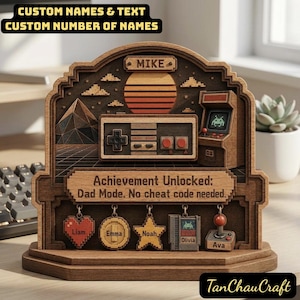 Puede incluir: Una pieza decorativa de madera con temática de juegos con el texto "CUSTOM NAMES & TEXT" y "CUSTOM NUMBER OF NAMES". Presenta un diseño retro de puesta de sol, un mando y una máquina recreativa. También está presente el texto "Achievement Unlocked: Dad Mode. No cheat code needed."
