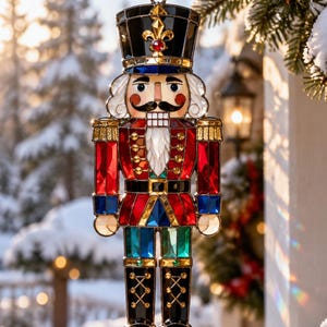 May include: A stained glass nutcracker ornament featuring a black hat with a gold fleur-de-lis, a red jacket with gold buttons, blue accents, and black boots. The figure has a white beard, a moustache, and a clear, multi-coloured glass construction.