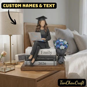 May include: A graduation figurine of a person in a cap and gown, holding a laptop, seated on books. The books are labelled "Emily", "Software Engineer", and "2026". A bouquet of blue and white flowers sits nearby. The text "CUSTOM NAMES & TEXT" is at the top.
