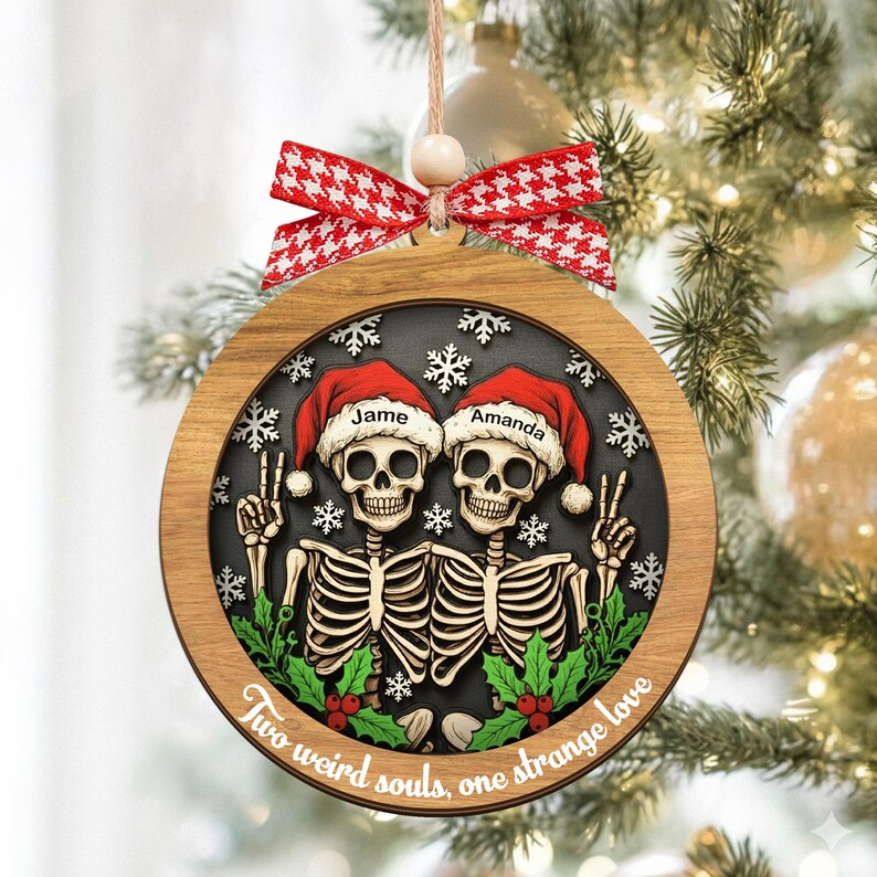 K&ouml;nnte beinhalten: Ein runder Weihnachtsbaumschmuck aus Holz mit zwei Skeletten in roten Weihnachtsm&uuml;tzen, beschriftet mit den Namen Jame und Amanda. Das Design zeigt wei&szlig;e Schneeflocken, gr&uuml;ne Stechpalmenbl&auml;tter mit roten Beeren und den Text Two weird souls, one strange love.