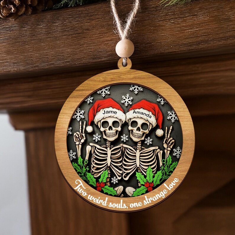K&ouml;nnte beinhalten: Ein runder Weihnachtsbaumschmuck aus Holz mit zwei Skeletten in roten Weihnachtsm&uuml;tzen, auf denen die Namen Jame und Amanda stehen. Die Skelette zeigen Friedenszeichen vor einem dunklen Hintergrund mit wei&szlig;en Schneeflocken, gr&uuml;nem Stechpalmenlaub und roten Beeren. Unten steht der Text Two weird souls, one strange love.
