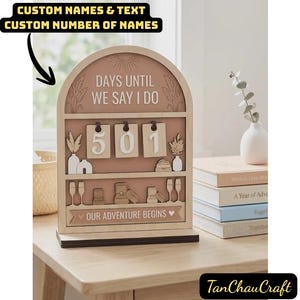 Puede incluir: Un calendario de cuenta regresiva de boda de madera con el texto "DAYS UNTIL WE SAY I DO" y "OUR ADVENTURE BEGINS". El calendario muestra el número "501" e incluye elementos decorativos como jarrones, copas de champán y anillos. También se ve el texto "CUSTOM NAMES & TEXT CUSTOM NUMBER OF NAMES".