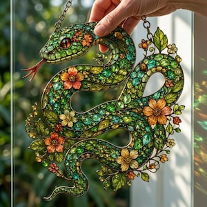 Könnte beinhalten: Ein Buntglas-Sonnenfänger in Form einer gewundenen Schlange mit grünem Körper, orangefarbenen und gelben Blumenakzenten und einer roten gespaltenen Zunge. Das Stück hat einen komplexen Metallrahmen und hängt an einer Metallkette.