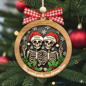 K&ouml;nnte beinhalten: Ein runder Weihnachtsbaumschmuck aus Holz mit zwei Skeletten in roten Weihnachtsm&uuml;tzen, auf denen die Namen Jame und Amanda stehen. Die Skelette zeigen das Friedenszeichen, umgeben von wei&szlig;en Schneeflocken und gr&uuml;nen Stechpalmenbl&auml;ttern. Unten steht der Text Two weird souls, one strange love, verziert mit einer rot-wei&szlig;en Hahnentritt-Schleife.