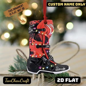 Könnte beinhalten: Ein dekorativer Weihnachtsschmuck in Form eines schwarz-roten Motocross-Stiefels mit dem Namen "Remi" an der Seite. Der Anhänger hat ein rotes Band zum Aufhängen und den Text "CUSTOM NAME ONLY" und "2D FLAT". Der Text "THIS ORNAMENT IS FLAT. NOT 3D" ist ebenfalls sichtbar.