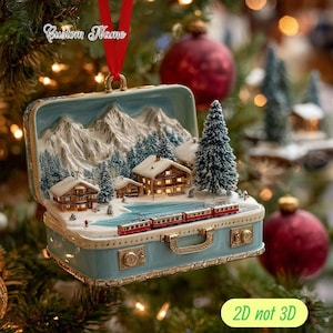 Pode incluir: Enfeite de Natal azul claro em forma de mala, apresentando uma cena de inverno com montanhas cobertas de neve, casas e um comboio vermelho. Possui uma fita vermelha para pendurar e o texto "Custom Name".