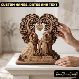 Könnte beinhalten: Eine herzförmige Holzskulptur mit zwei Paaren, einem jüngeren und einem älteren, die unter einem Baum stehen. Ein kleiner Herzanhänger hängt in der Mitte mit dem Text 'Then, Now, Always'. Der Sockel ist mit 'Sarah', 'David' und 'Then, Now, Always' graviert.