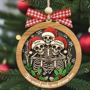 K&ouml;nnte beinhalten: Ein runder Weihnachtsbaumschmuck aus Holz mit drei Skeletten in roten Weihnachtsm&uuml;tzen. Die M&uuml;tzen sind mit den Namen Jame und Amanda personalisiert. Die Skelette sind von gr&uuml;nen Stechpalmenbl&auml;ttern und wei&szlig;en Schneeflocken umgeben, mit dem Text Two weird souls, one strange love. Eine rot-wei&szlig; gemusterte Schleife ist oben befestigt.