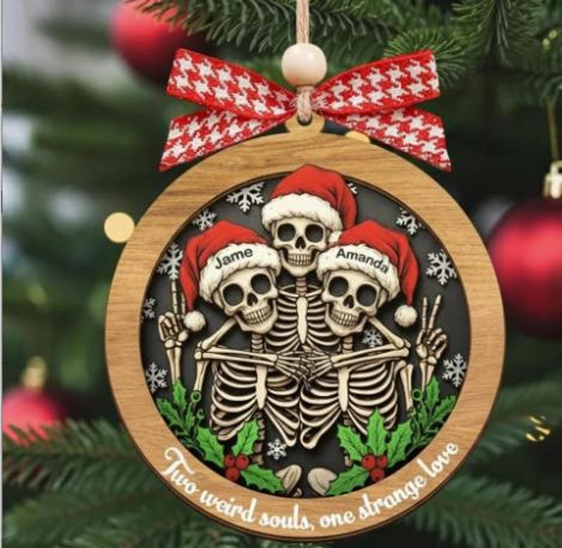 K&ouml;nnte beinhalten: Ein runder Weihnachtsbaumschmuck aus Holz mit drei Skeletten in roten Weihnachtsm&uuml;tzen. Die M&uuml;tzen sind mit den Namen Jame und Amanda personalisiert. Die Skelette sind von gr&uuml;nen Stechpalmenbl&auml;ttern und wei&szlig;en Schneeflocken umgeben, mit dem Text Two weird souls, one strange love. Eine rot-wei&szlig; gemusterte Schleife ist oben befestigt.