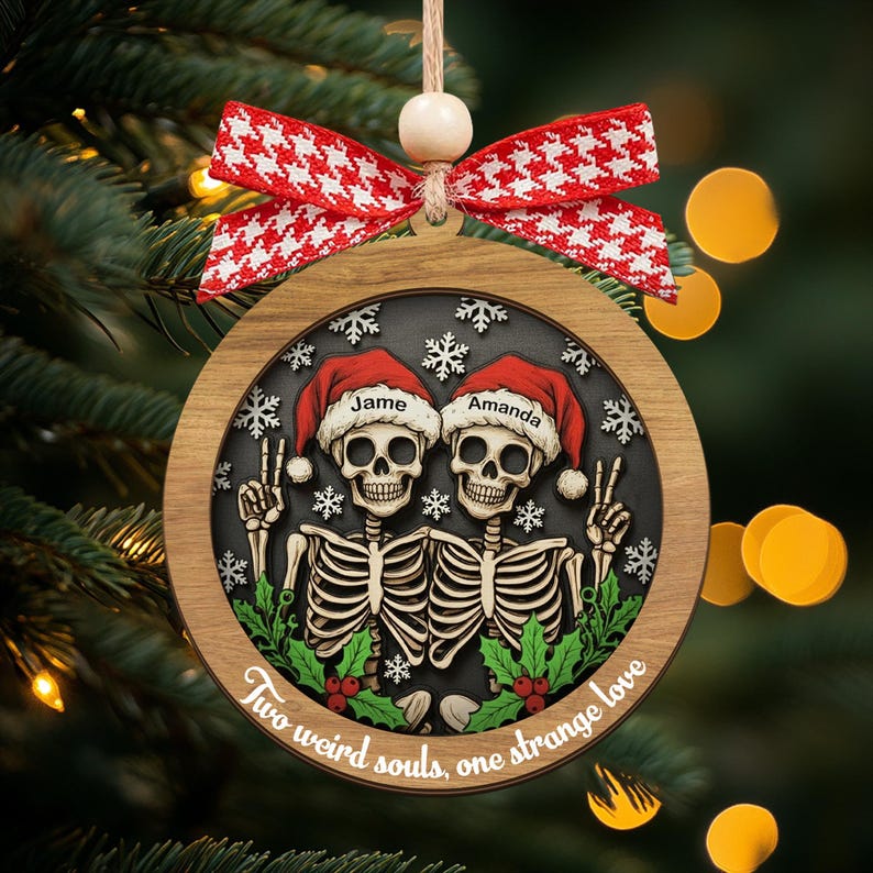 K&ouml;nnte beinhalten: Ein runder Weihnachtsbaumschmuck aus Holz mit zwei Skeletten in roten Weihnachtsm&uuml;tzen, auf denen die Namen Jame und Amanda stehen. Die Skelette zeigen das Friedenszeichen, umgeben von wei&szlig;en Schneeflocken, gr&uuml;nem Stechpalmenlaub und dem Text Two weird souls, one strange love. Oben ist eine rot-wei&szlig;e Hahnentritt-Schleife befestigt.