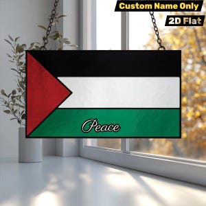 Può includere: Un cartello rettangolare con il design della bandiera palestinese, con colori nero, bianco, verde e rosso. La parola "Peace" è scritta in corsivo in basso. Il cartello è appeso a catene.