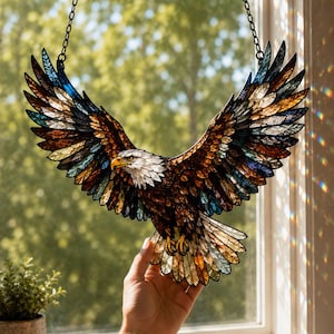 Könnte beinhalten: Ein Buntglas-Sonnenfänger in Form eines Weißkopfseeadlers im Flug mit ausgebreiteten Flügeln. Der Adler hat einen weißen Kopf, einen gelben Schnabel und ein Gefieder aus braunen, bernsteinfarbenen und blauen Glasstücken, aufgehängt an einer Kette.