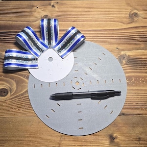 May include: A craft project in progress, featuring a gray circular base with evenly spaced notches, a white circular piece, and a black pen. A decorative bow with blue, black, and silver striped ribbon is attached to the white circle.