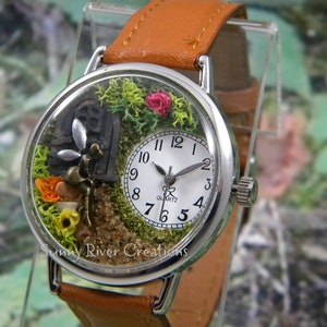 May include: A silver wristwatch with a brown leather band. The watch face is encased in a clear dome with a miniature fairy garden scene inside. The scene includes a miniature fairy, a small house, moss, flowers, and sand.