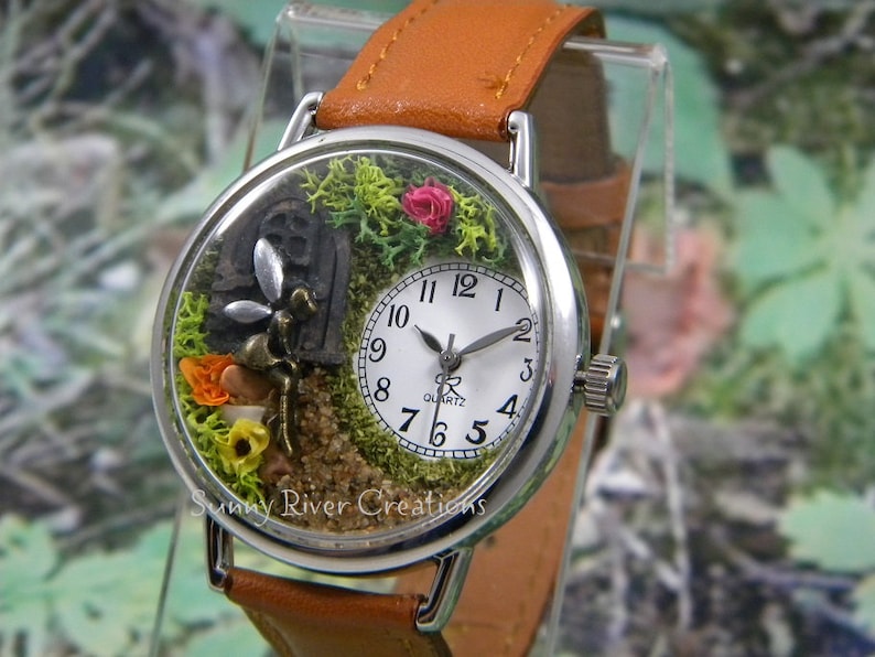 May include: A silver wristwatch with a brown leather band. The watch face is encased in a clear dome with a miniature fairy garden scene inside. The scene includes a miniature fairy, a small house, moss, flowers, and sand.