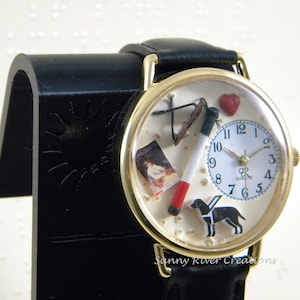 May include: A gold-tone wristwatch with a black leather strap featuring a cream-colored dial. The dial includes a small clock face, a miniature book, a cigarette, a red heart, and a black dog figure. The text Sunny River Creations is visible.