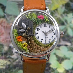 May include: A silver wristwatch with a white face and black hands. The watch face is encased in a clear dome with a miniature fairy garden inside. The garden features moss, flowers, and a small fairy figurine.