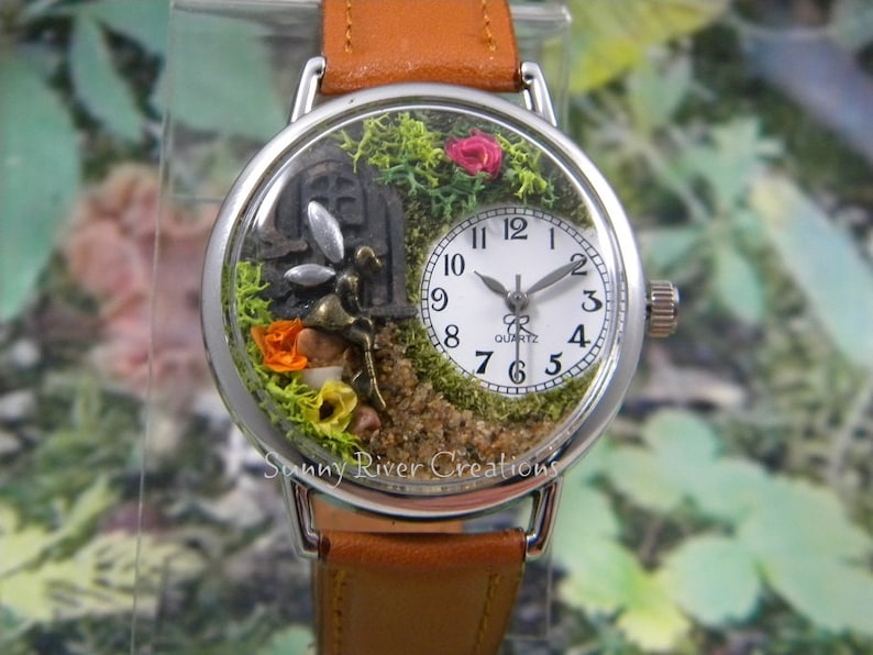 May include: A silver wristwatch with a white face and black hands. The watch face is encased in a clear dome with a miniature fairy garden inside. The garden features moss, flowers, and a small fairy figurine.