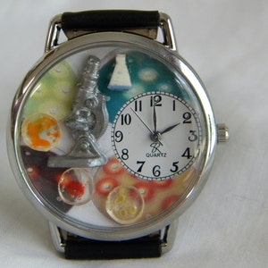 May include: A silver-toned analog wristwatch with a black leather strap. The watch face features a white dial with black numerals and the text 'QUARTZ'. The background displays a silver microscope, a white flask, and colorful circular petri dish motifs.