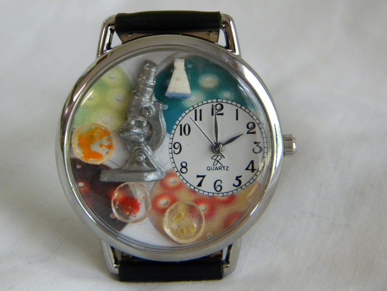 May include: A silver-toned analog wristwatch with a black leather strap. The watch face features a white dial with black numerals and the text 'QUARTZ'. The background displays a silver microscope, a white flask, and colorful circular petri dish motifs.