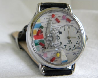 Puede incluir: Reloj de cuarzo plateado con correa de cuero negro. La esfera presenta un microscopio, tubos de ensayo en miniatura y un diagrama. Las manecillas señalan las 2:10. Un accesorio único con temática médica o científica.