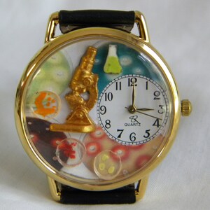 May include: A gold-tone wristwatch with a black leather strap. The watch face features a gold microscope, a small green flask, and colorful petri dish illustrations. A white circular dial with black Arabic numerals displays the time, marked Quartz.