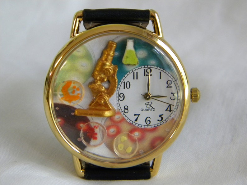 May include: A gold-tone wristwatch with a black leather strap. The watch face features a gold microscope, a small green flask, and colorful petri dish illustrations. A white circular dial with black Arabic numerals displays the time, marked Quartz.