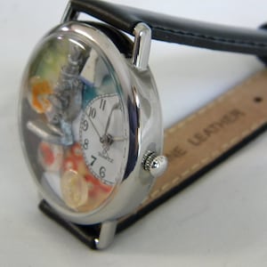May include: A silver-tone wristwatch with a black leather strap. The watch face features a white dial with black numerals, a silver microscope charm, and colorful decorative elements inside the clear casing. The word QUARTZ is printed on the dial.