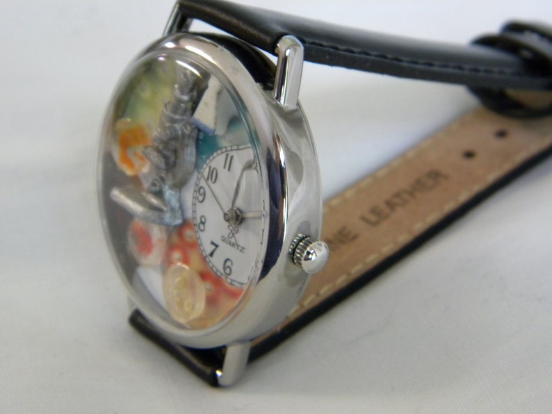May include: A silver-tone wristwatch with a black leather strap. The watch face features a white dial with black numerals, a silver microscope charm, and colorful decorative elements inside the clear casing. The word QUARTZ is printed on the dial.