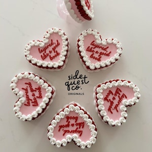 Pode incluir: Biscoitos em forma de coração com cobertura rosa e borda de creme branco. Os biscoitos têm as palavras "layered, like a good book" escritas em vermelho. Os biscoitos são dispostos em torno de um biscoito central com um suporte transparente.