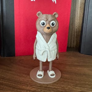 May include: A brown bear figurine standing on a circular base, wearing a white bathrobe and white slippers. The bear has large, wide eyes and a serious expression. Behind the figurine is a red book cover featuring a white anarchy symbol.