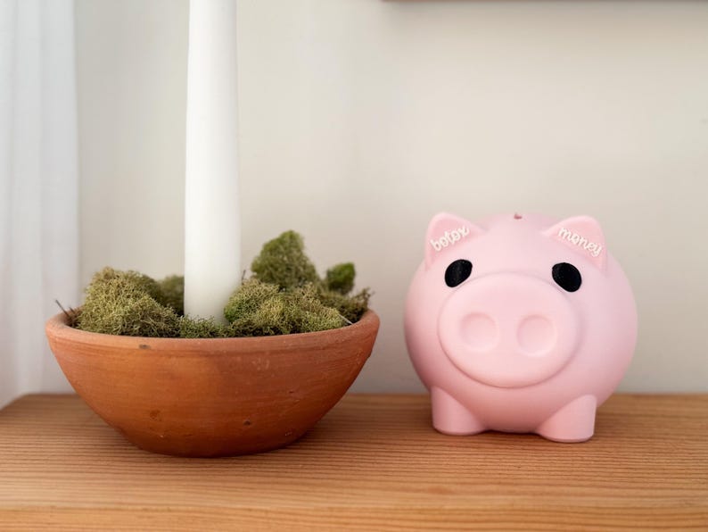 May include: A pink piggy bank with the words "baby" and "money" on its ears sits on a wooden surface. Next to it is a terracotta bowl filled with green moss and a white candle.