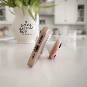 May include: A tan phone case with a white pop-up grip featuring a pink and brown design. The phone case is resting on a white surface. A white vase with the text "side quest co. originals" is in the background.