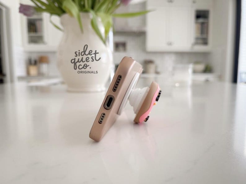May include: A tan phone case with a white pop-up grip featuring a pink and brown design. The phone case is resting on a white surface. A white vase with the text "side quest co. originals" is in the background.