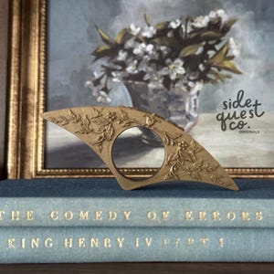 May include: A gold-coloured metal ring with a floral design and a circular opening. The ring is resting on top of two blue books with gold lettering. The background includes a framed painting and the text "side quest co. originals."