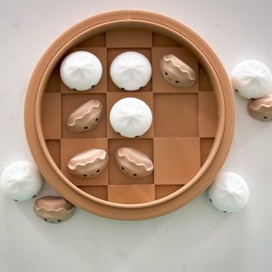 May include: A brown wooden board game with a chequered pattern. The board game is filled with small white and brown dumplings with faces. There are also several dumplings outside of the board game.