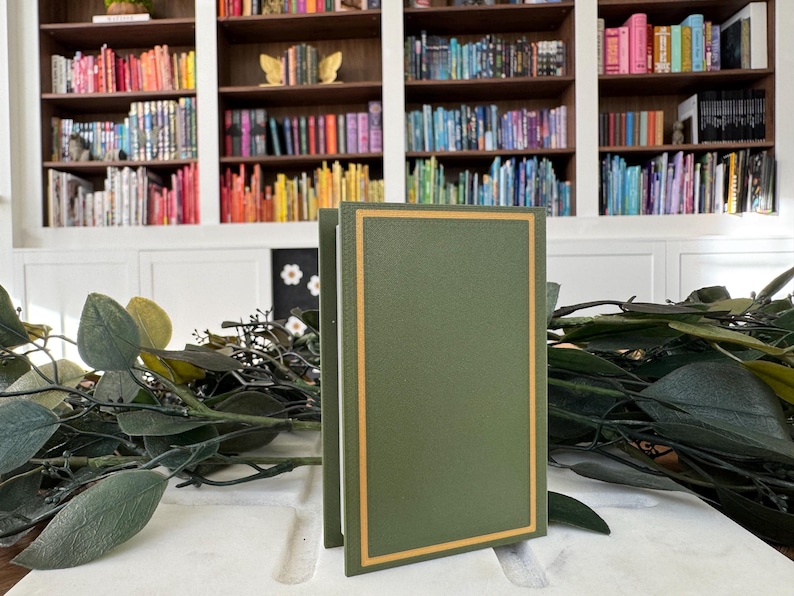 Puede incluir: Dos libros verdes con borde dorado se alzan sobre un fondo de una estanter&iacute;a llena de libros coloridos. La vegetaci&oacute;n y las hojas est&aacute;n en primer plano. Los libros tienen un dise&ntilde;o cl&aacute;sico y elegante.