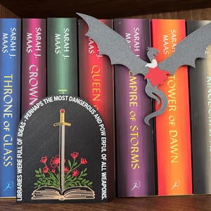 Könnte beinhalten: Ein schwarzer und goldener Buchstütze mit einem Schwert, einem Buch und roten Blumen. Die Buchstütze ist bogenförmig und trägt den Text "Bibliotheken waren voller Ideen - vielleicht die gefährlichsten und mächtigsten Waffen von allen." Die Buchstütze steht vor einem Bücherregal mit einem Drachen-Buchstütze und einer Reihe von Büchern von Sarah J. Maas.