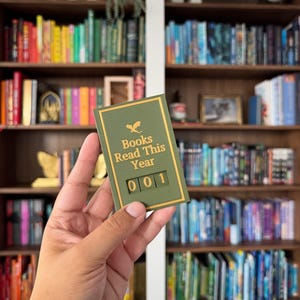 Puede incluir: Un contador de libros verde y dorado con el texto "Books Read This Year" y el n&uacute;mero "001". El contador se sostiene frente a una estanter&iacute;a llena de libros coloridos.