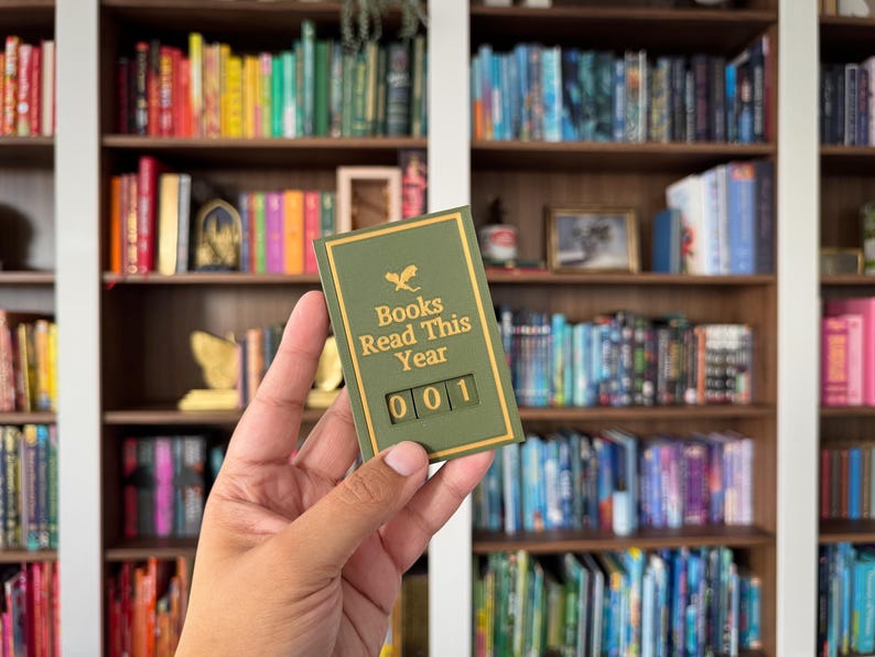 Puede incluir: Un contador de libros verde y dorado con el texto "Books Read This Year" y el n&uacute;mero "001". El contador se sostiene frente a una estanter&iacute;a llena de libros coloridos.
