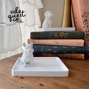 May include: A decorative tray with a small, speckled, light gray finish, holding a small, light gray French bulldog figurine. The tray is on a wooden surface, with a stack of vintage books in the background.
