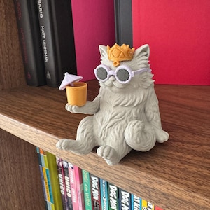 May include: A light gray cat figurine wearing a gold crown and round lavender sunglasses. The cat is sitting and holding a small orange cup with a tiny white umbrella. The figure has textured fur and is positioned on a wooden shelf in front of books.