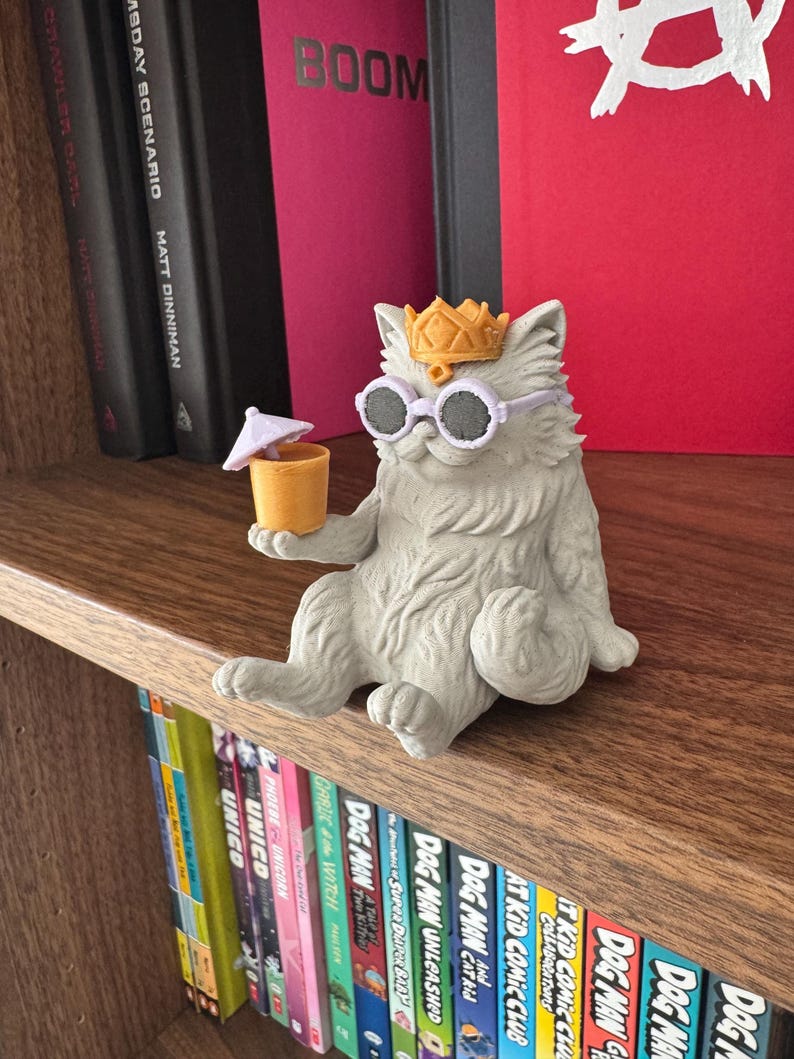 May include: A light gray cat figurine wearing a gold crown and round lavender sunglasses. The cat is sitting and holding a small orange cup with a tiny white umbrella. The figure has textured fur and is positioned on a wooden shelf in front of books.