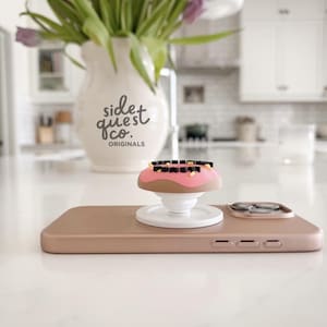 May include: A rose gold phone with a donut-shaped phone grip. The grip has pink frosting, brown donut, and black and yellow sprinkles. A white vase with the text "side quest co. originals" and purple flowers is in the background.
