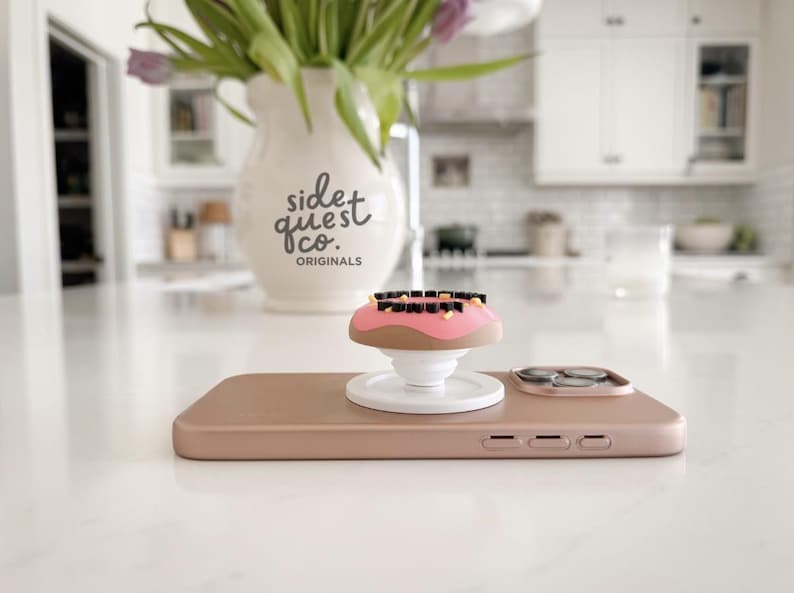 May include: A rose gold phone with a donut-shaped phone grip. The grip has pink frosting, brown donut, and black and yellow sprinkles. A white vase with the text "side quest co. originals" and purple flowers is in the background.