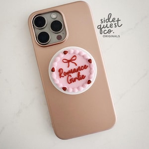 May include: A rose gold phone with a decorative pop socket. The pop socket is designed to look like a pink cake with the words "Romance Girlie" in red script. The cake has white frosting and red strawberry accents.