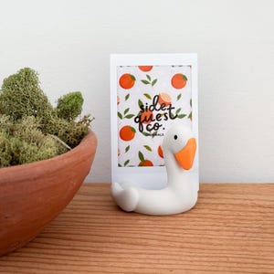 Könnte beinhalten: Ein weißer, gänseförmiger Bilderrahmen mit orangefarbenem Schnabel und Füßen. Der Rahmen enthält einen Druck mit Orangen und grünen Blättern. Der Text "side quest co. originals" ist sichtbar. Der Rahmen steht auf einer Holzoberfläche.